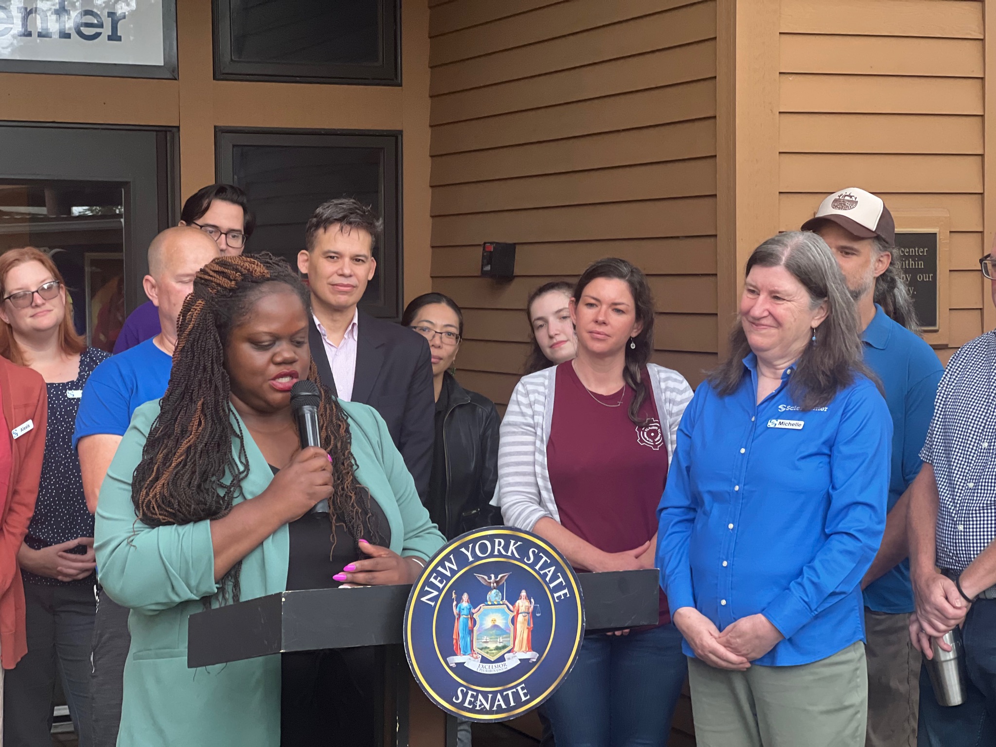 A black woman is holding a microphone behind a podium with a New York State Senate emblem. A group of adults is listening adjacent to and behind her. 